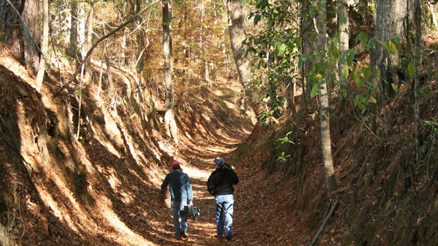 2 hikers walk away from viewer. 20 foot steep embankments are on both sides of the trail.